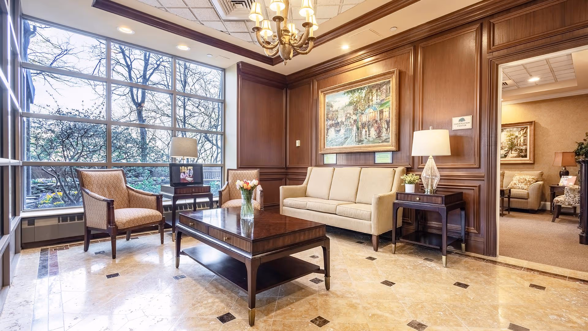 A bright and elegant living room area with large floor-to-ceiling windows showing trees outside. The room features a beige sofa, two upholstered armchairs, a dark wooden coffee table with a vase of flowers, and side tables with lamps. The walls are paneled with rich wood, and a chandelier hangs from the ceiling. A painting is mounted on the wall above the sofa, and an adjacent room with additional seating is visible through an open doorway.