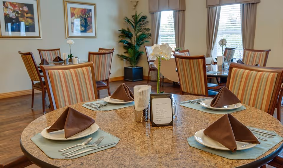 A dining room with round tables set for four people each, featuring plates with folded brown napkins, silverware, and light blue placemats. The room has striped upholstered chairs, framed artwork on the walls, large windows with beige curtains, and a potted plant in the corner.