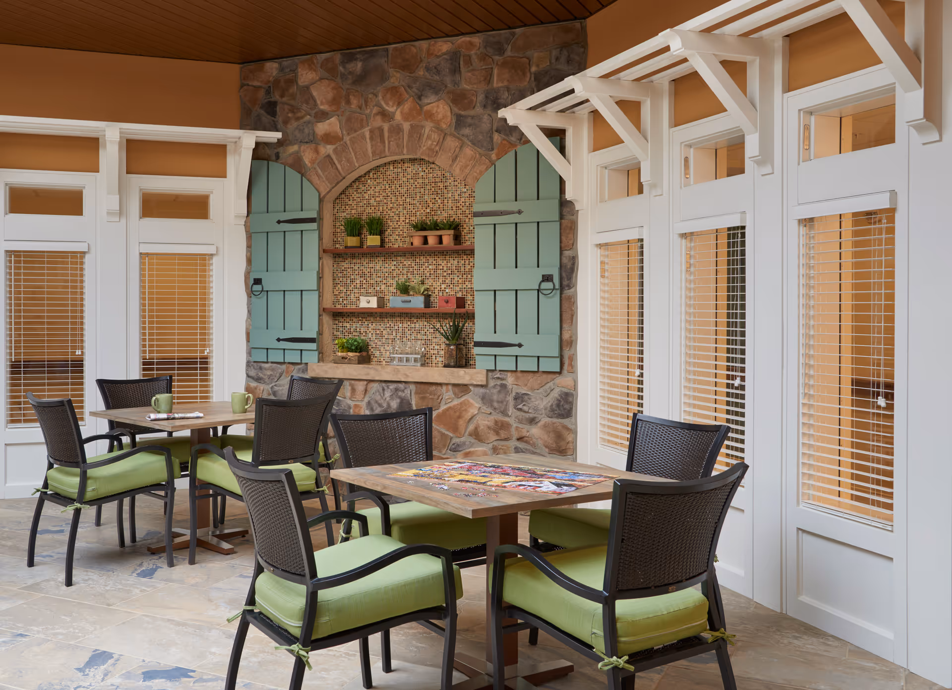 Sunlit communal seating area with tables, green-cushioned chairs, and a stone alcove with decorative shutters and shelves.