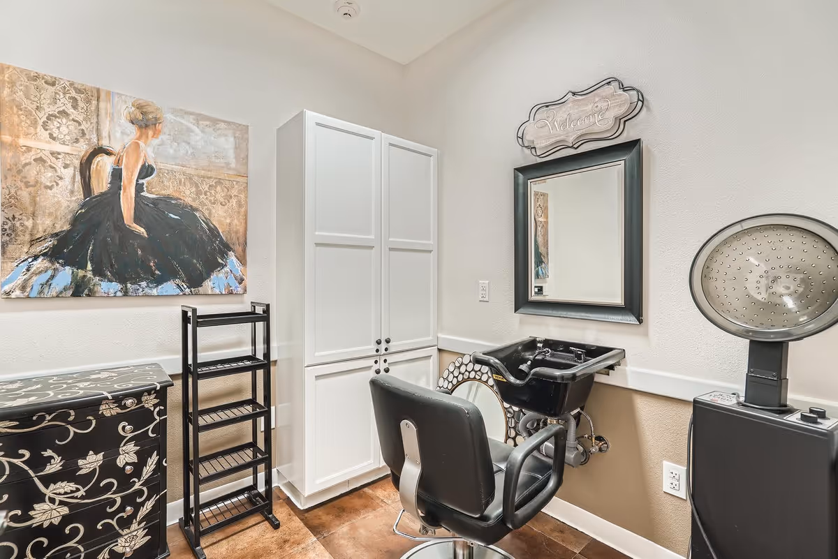 Interior view of a salon area in a senior living facility featuring a black salon chair in front of a black wash basin, a large mirror with a decorative 'Welcome' sign above it, a black hair dryer, white storage cabinets, a black shelving unit, and a painting of a woman in a black dress sitting on a chair on the wall.