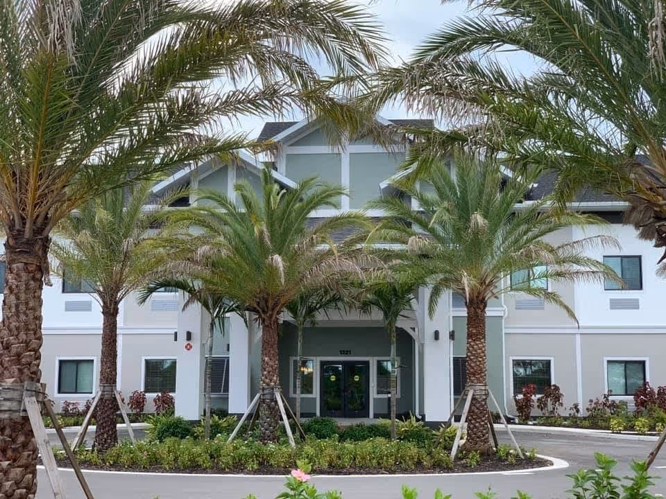 Front exterior view of a two-story senior living facility building with a light green and white facade, surrounded by palm trees and landscaped greenery under a cloudy sky.