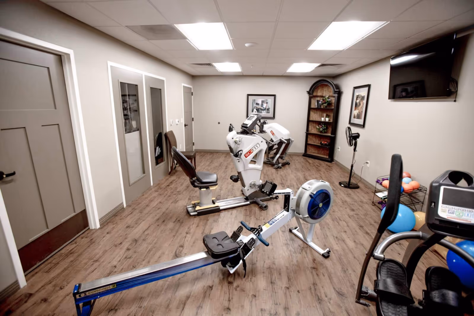 A fitness room with exercise equipment including a rowing machine, a recumbent bike, and other workout machines. The room has wood flooring, beige walls, a wall-mounted TV, framed pictures, a tall wooden shelf with plants and decorations, and a small fan. There are three closed doors along one wall.