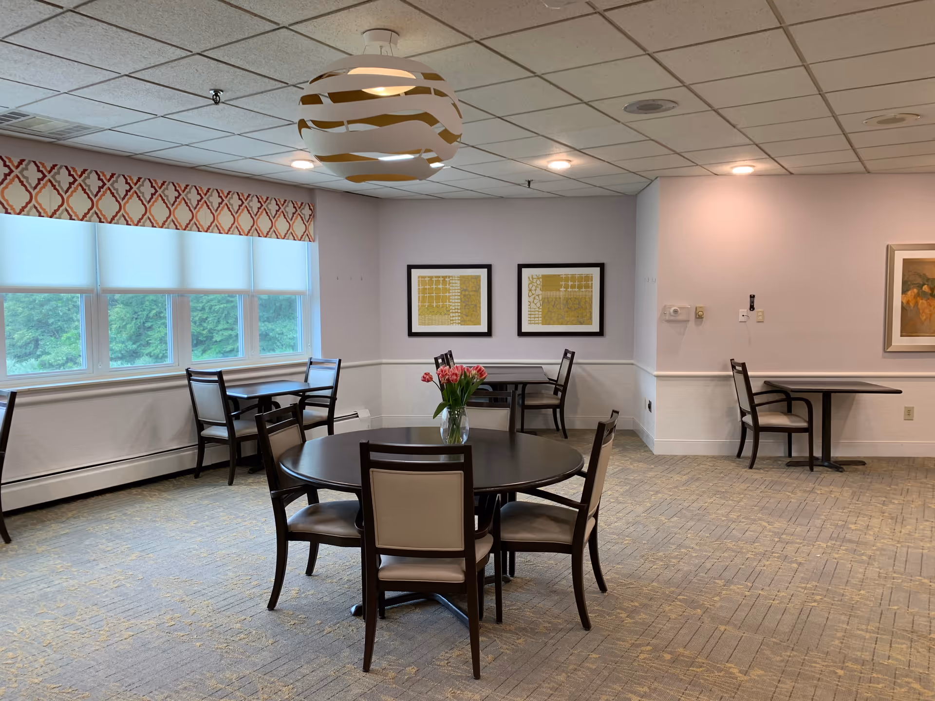 A dining room with several tables and chairs arranged around the room. A round table with a vase of pink tulips is in the foreground. Large windows with white blinds and patterned valances let in natural light. The walls are decorated with framed artwork, and a modern white and gold pendant light hangs from the ceiling.