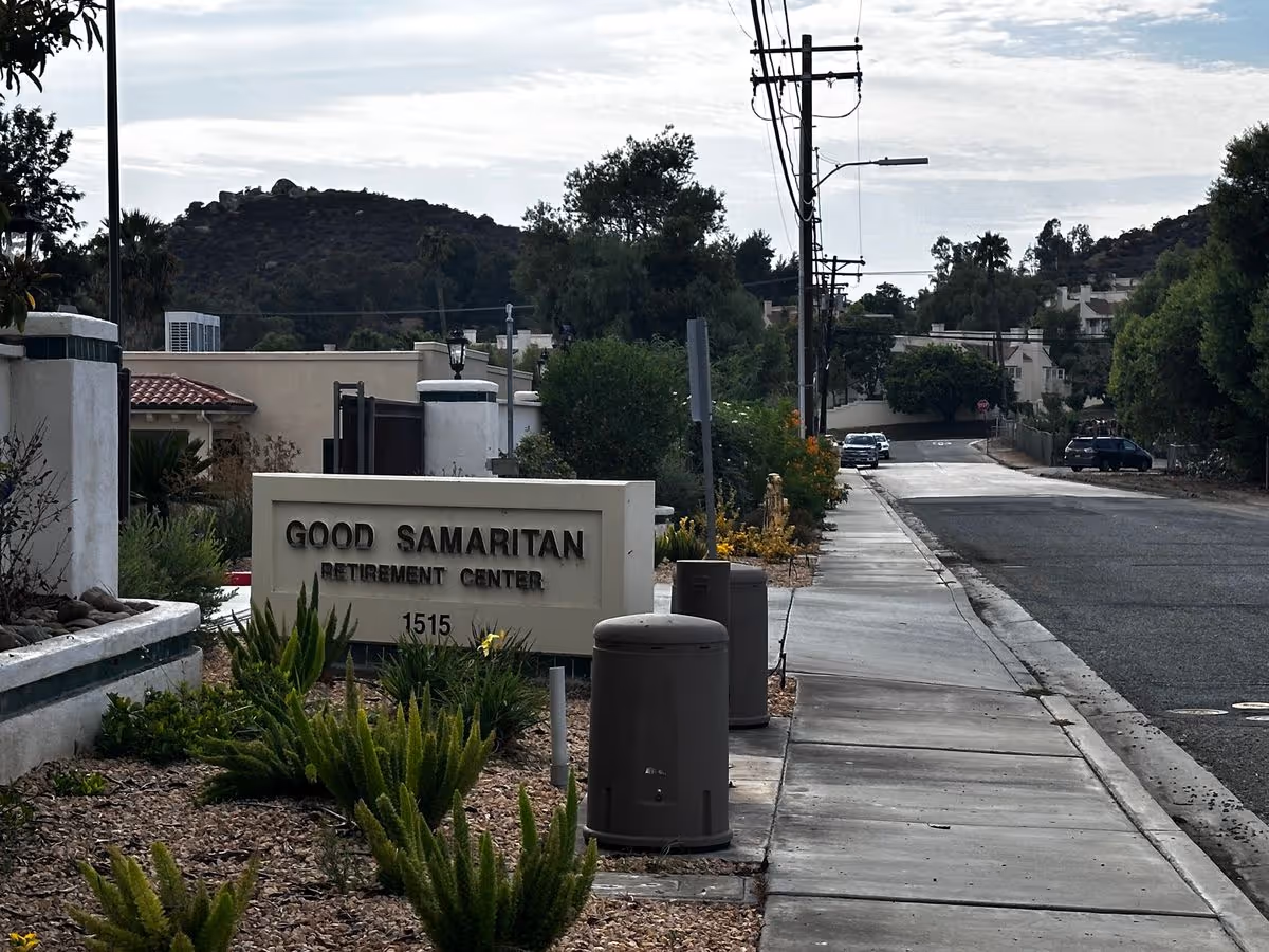 Entrance sign for Good Samaritan Retirement Center next to a sidewalk and street with landscaping and distant hills.