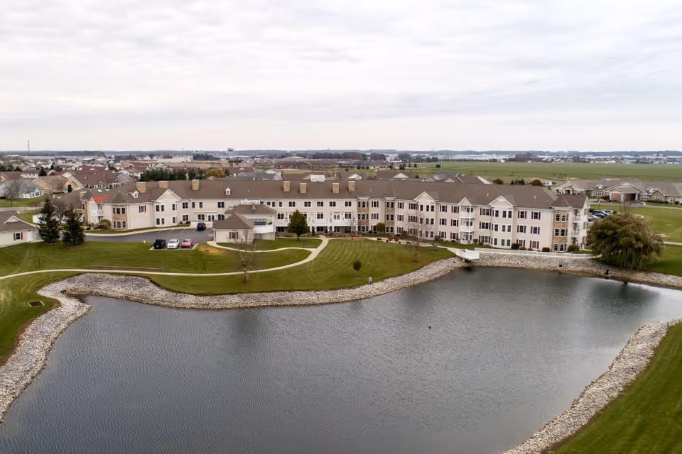 Aerial view of Waterford Crossing senior living facility showing a large multi-story building with beige siding and brown roofs situated next to a pond with a stone-lined edge. The surrounding area includes green lawns, walking paths, a few parked cars, and other residential buildings in the background under a cloudy sky.