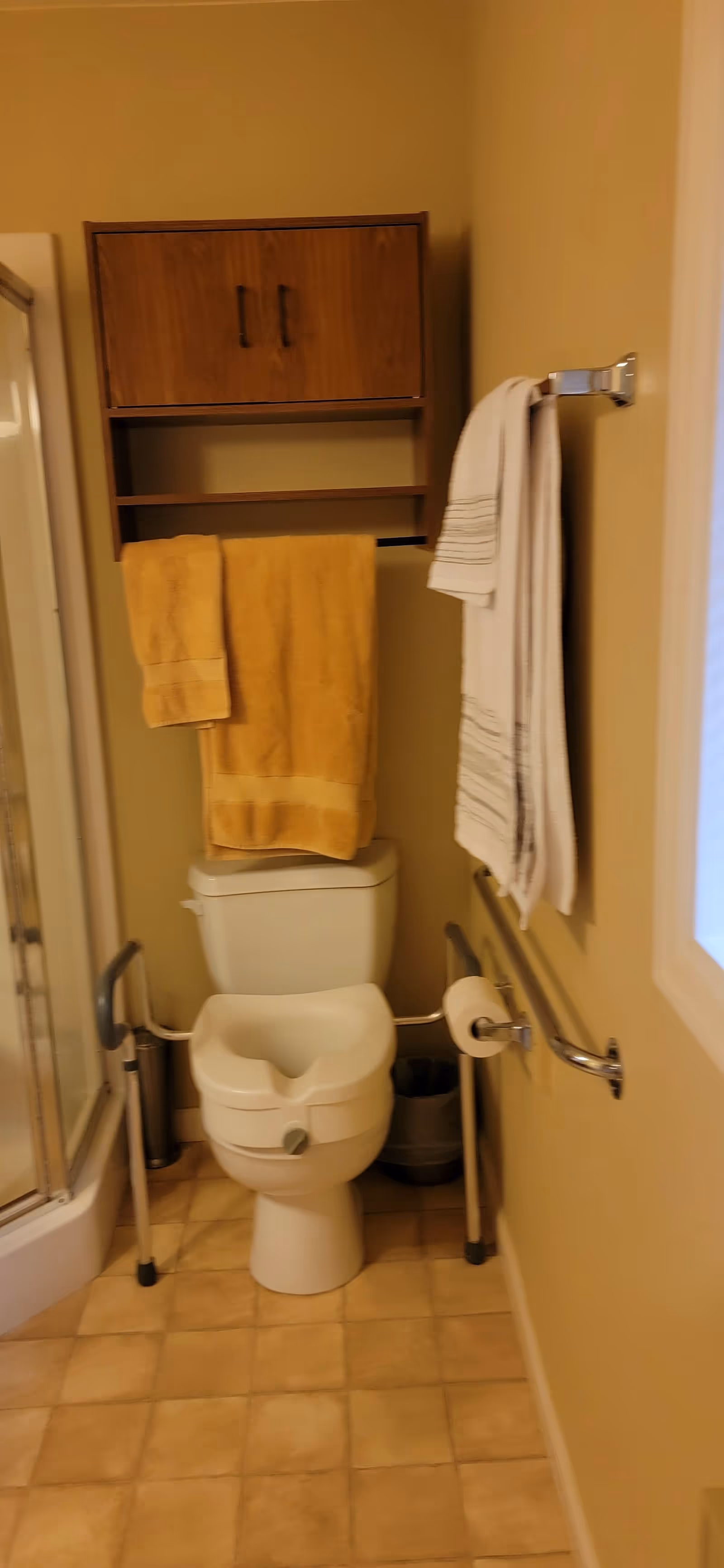 Bathroom with a toilet equipped with safety rails and a raised seat. Above the toilet is a wooden cabinet with two doors and an open shelf. Two towels, one yellow and one white with stripes, hang on towel racks. The floor has beige tiles and there is a shower stall visible on the left side.