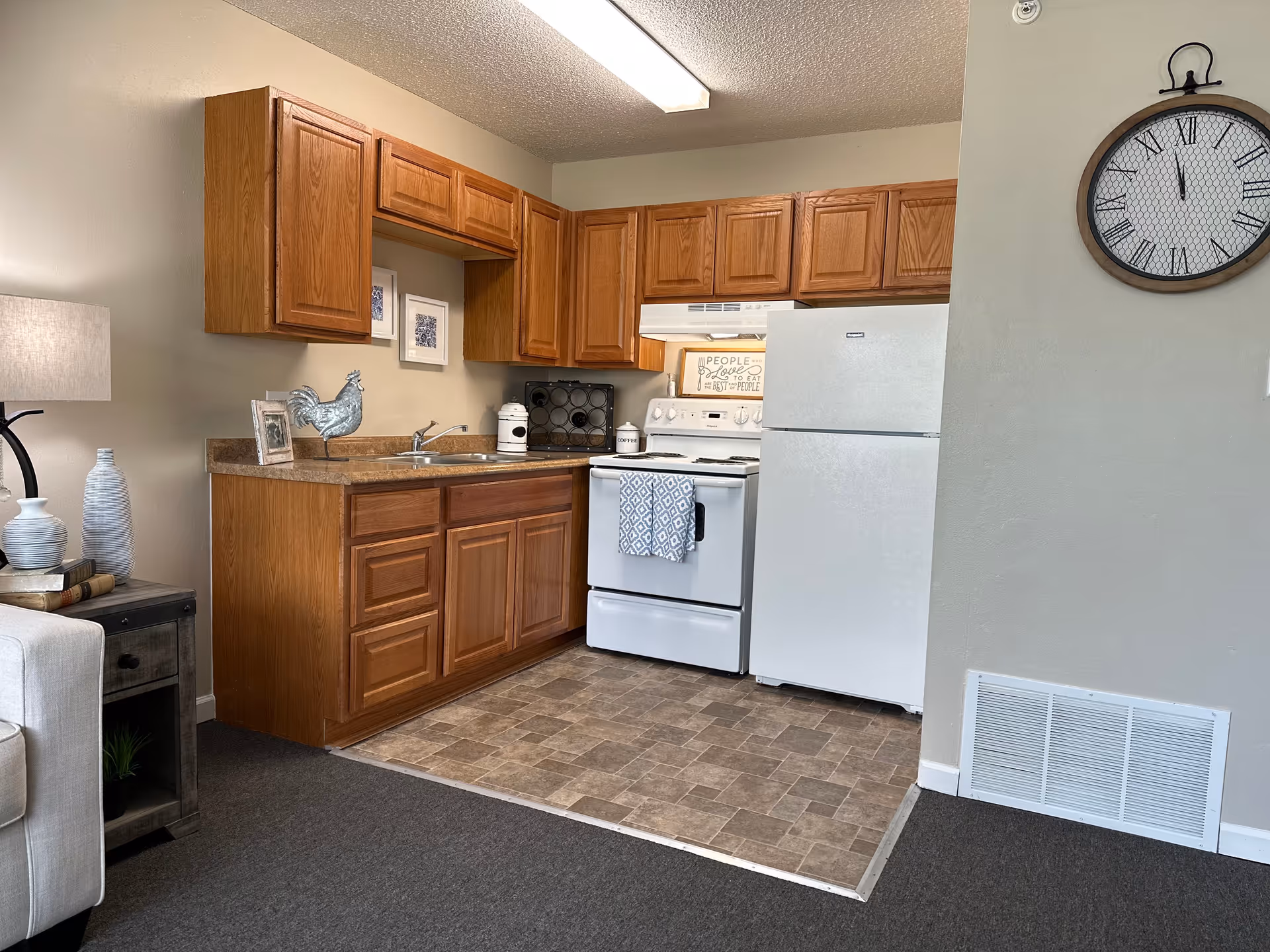 A small kitchen area with wooden cabinets, a white refrigerator, and a white stove with a patterned towel hanging on the oven door. The countertop has a sink, a metal rooster decoration, a coffee canister, and a wine rack. To the left is a side table with a lamp, books, and decorative vases. A large round wall clock with Roman numerals hangs on the wall to the right.