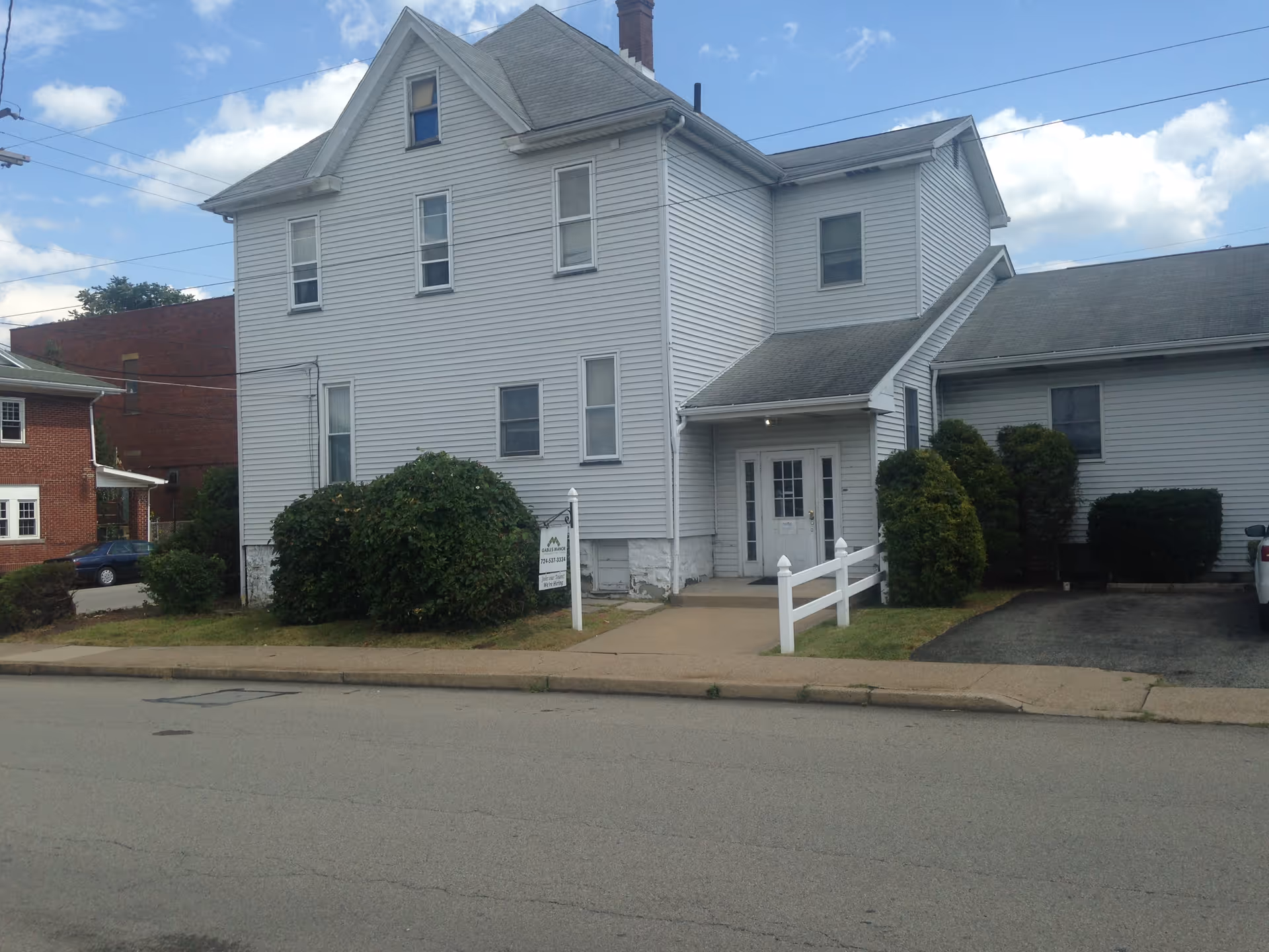 White two-story building with a small porch, sidewalk, and shrubs along a street.