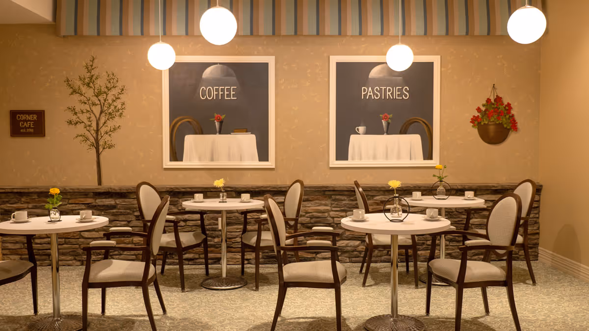 A cozy cafe area with round tables and cushioned chairs arranged neatly. Each table has a small vase with a yellow flower and white coffee cups. The wall features two framed pictures labeled 'COFFEE' and 'PASTRIES' above the tables. There is a small sign on the left wall that reads 'CORNER CAFE est. 2016' and a hanging basket with red flowers on the right. Three round pendant lights hang from the ceiling, illuminating the space warmly.