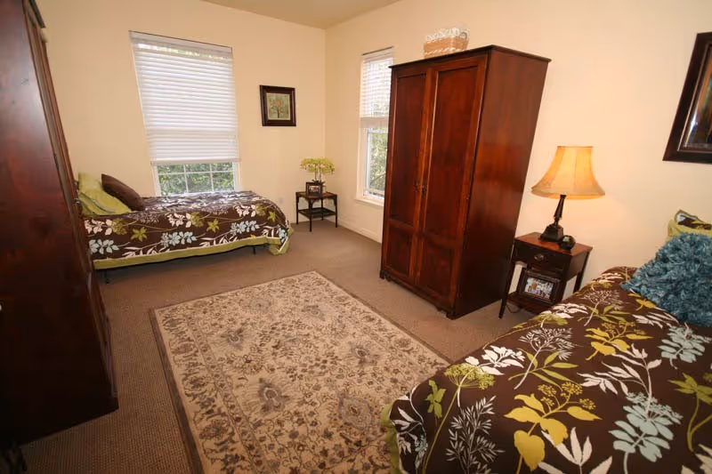 A bedroom with two twin beds featuring brown floral bedspreads, a wooden wardrobe, a nightstand with a lamp, a small table with a plant, two windows with blinds, and a beige patterned rug on the floor.