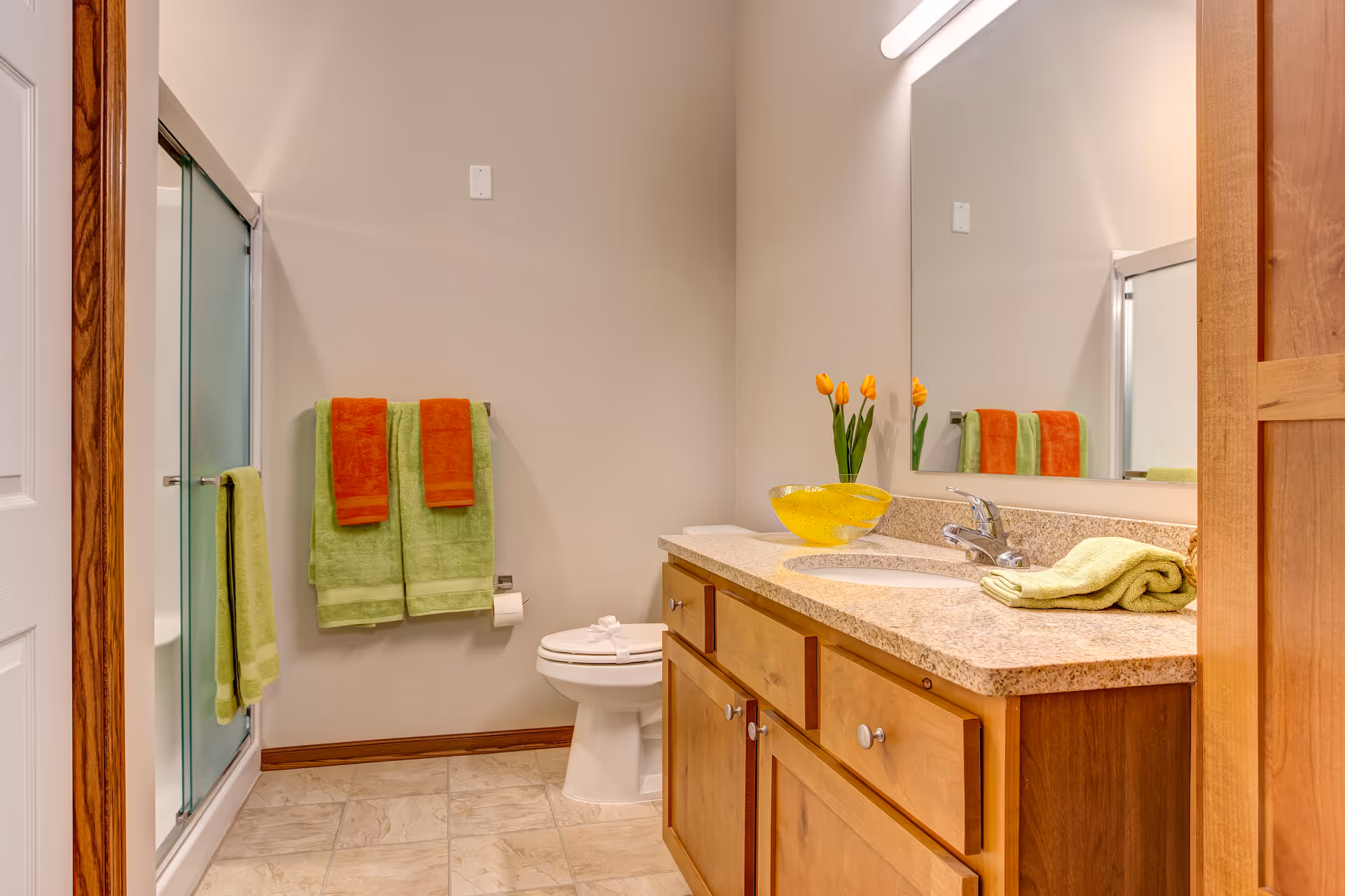 A clean and well-lit bathroom with a wooden vanity topped with a granite countertop, a sink, and a large mirror above it. There are yellow tulips in a glass vase and a yellow decorative bowl on the countertop. On the wall opposite the vanity, there is a toilet and a towel rack holding green and orange towels. A glass-enclosed shower is visible to the left.