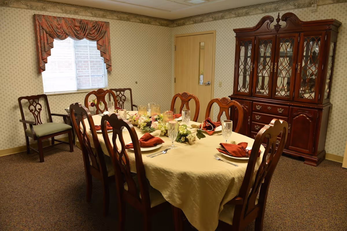 A formal dining room with a long rectangular table covered with a beige tablecloth. The table is set with plates, silverware, crystal glasses, and red cloth napkins. A floral centerpiece runs down the middle of the table. Surrounding the table are wooden chairs with intricate backs. Against the wall is a large wooden china cabinet with glass doors displaying glassware. A window with a red valance and a wooden chair are also visible. The walls have a subtle patterned wallpaper.