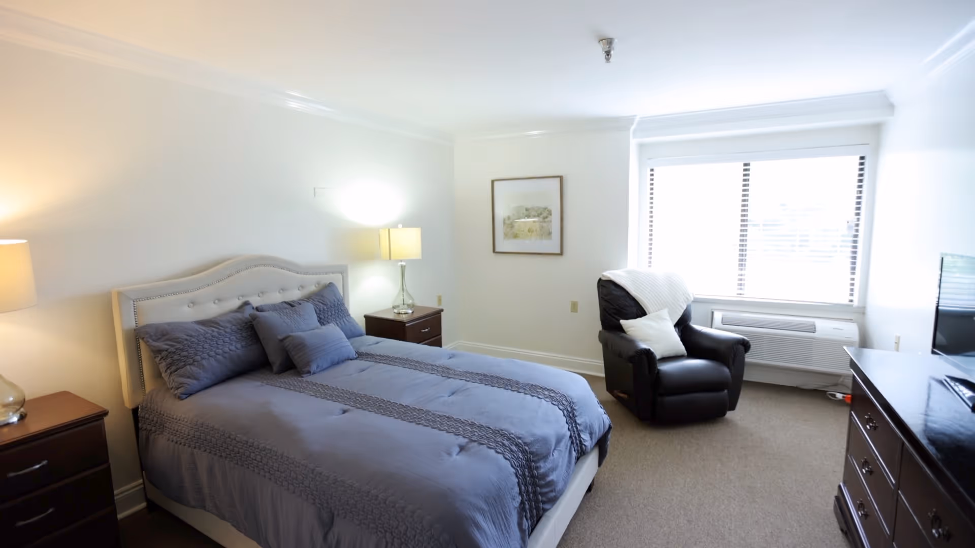 Bright, neatly furnished bedroom with a made bed, two nightstands and lamps, a recliner by a large window, and a dresser with a TV.