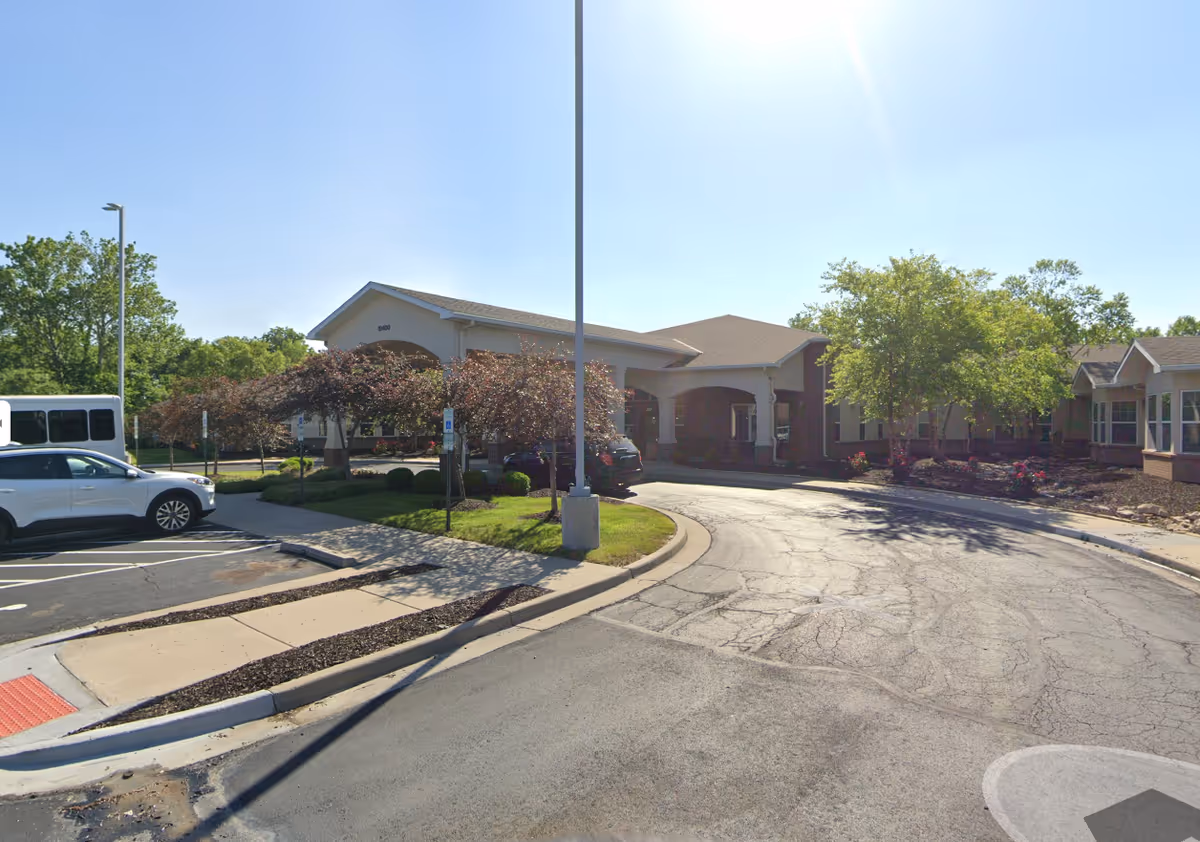 Exterior view of a single-story building with a covered entrance driveway, surrounded by trees and parked cars under a clear blue sky.