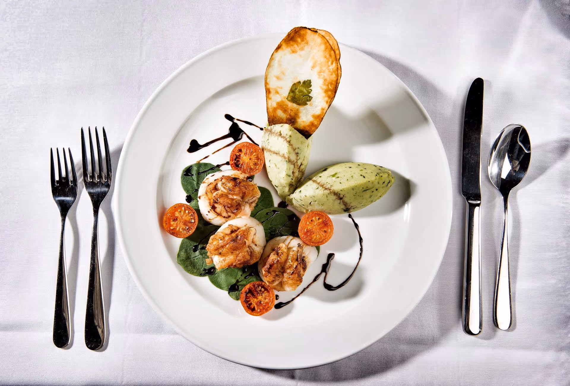 A white plate with a gourmet dish consisting of three seared scallops on a bed of spinach leaves, garnished with halved cherry tomatoes, two quenelles of green herb butter or mousse, a crisp chip with a small leaf on top, and a drizzle of balsamic reduction. The plate is set on a white tablecloth with two forks on the left and a knife and spoon on the right.