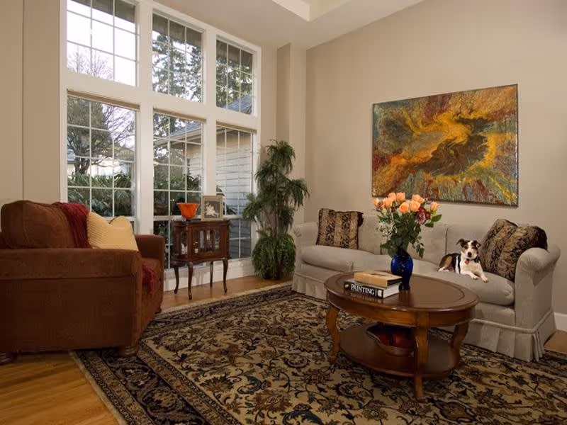 A cozy living room with a large window letting in natural light. The room features a brown armchair with a yellow pillow and red throw, a beige sofa with patterned cushions, and a small dog sitting on the sofa. A wooden coffee table with a vase of flowers and books sits on an ornate patterned rug. There is a tall green plant near the window and a colorful abstract painting on the wall.