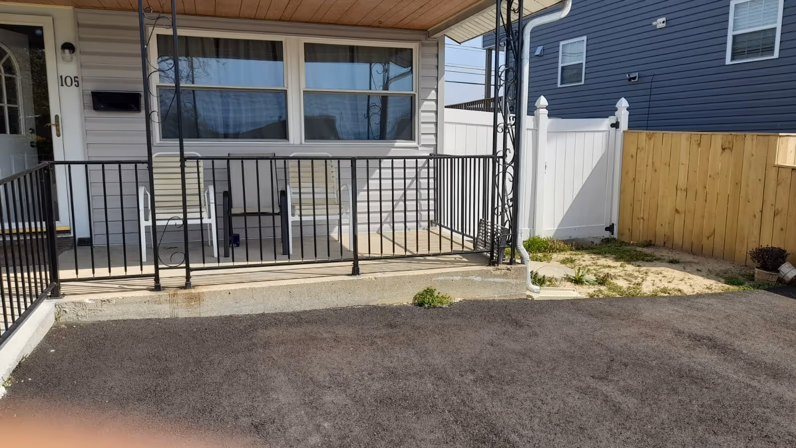 Front porch and entrance of a single-story unit with two chairs behind a metal railing, a door labeled '105', and a paved area in front.