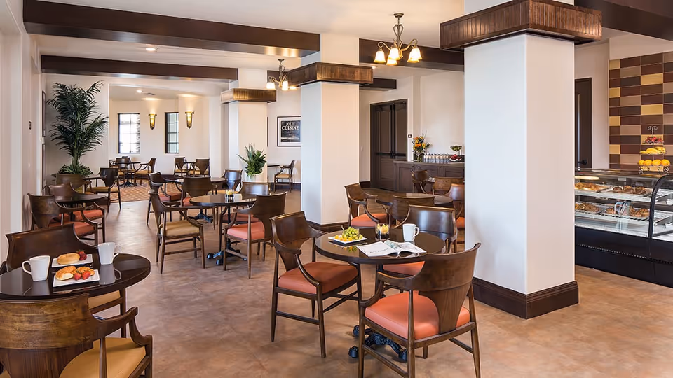 A bright and spacious dining area with multiple round wooden tables and chairs with orange cushions. The room features white walls with dark wooden beams and trim, potted plants, and a display case filled with pastries on the right side. There are plates with food and cups on some tables, and the overall ambiance is warm and inviting.