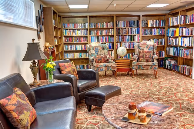 A cozy library room with wooden bookshelves filled with books lining the walls. The room features two patterned armchairs with a small table and globe between them, a black leather sofa and armchair with floral cushions, a matching ottoman, a table lamp, and a round marble coffee table with decorative items and a book on it. The floor is carpeted with a patterned design, and a window with blinds allows natural light into the space.
