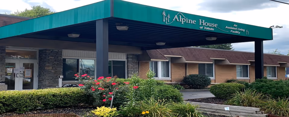Exterior view of Alpine House of Toledo, an assisted living facility, showing the entrance with a green canopy, stone and brick walls, windows, and landscaped bushes and flowers in front.