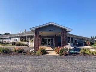 Front exterior view of a single-story senior living facility building with a covered entrance supported by brick columns, surrounded by landscaped bushes and flowers under a clear blue sky.