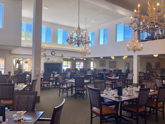 Spacious dining room with multiple tables and chairs arranged neatly. The room features high ceilings with large windows letting in natural light, and elegant chandeliers hanging from the ceiling. Tables are set with cups, glasses, and napkins, ready for guests. A fireplace is visible in the background along with some wall decorations.