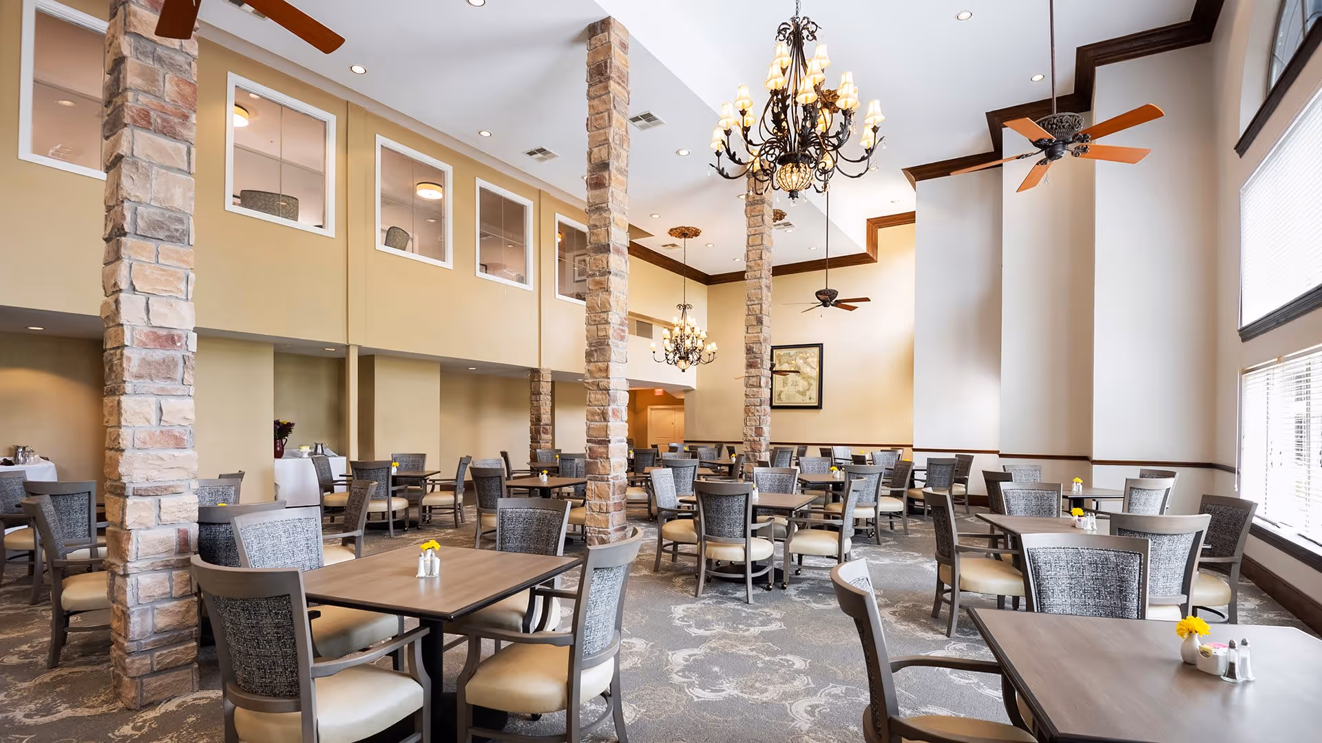 A spacious dining room with multiple tables and chairs arranged neatly. The room features tall ceilings with stone pillars, ceiling fans, and elegant chandeliers. Large windows on one side allow natural light to fill the space. Each table has a small flower vase and salt and pepper shakers.