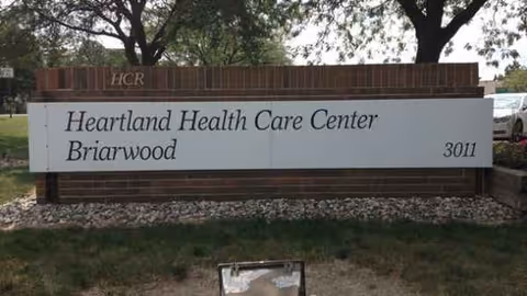 A brick sign for Heartland Health Care Center Briarwood sits on landscaped grounds.