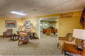 Interior view of a senior living facility lounge area named Trinity Chapel. The room features patterned carpet, several upholstered chairs, a small round table with a lamp, framed artwork on the walls, and a doorway leading to another room with additional seating.