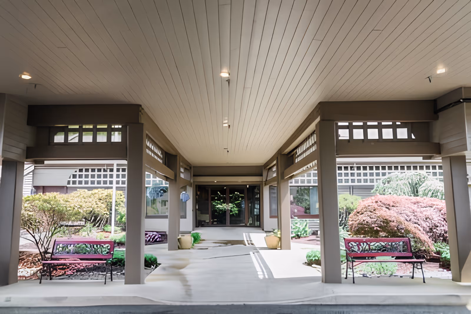 Covered outdoor walkway with benches on either side and landscaped bushes and plants, leading to a building entrance with glass doors at Channel Point Village.
