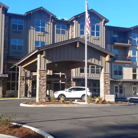 Exterior view of a multi-story senior living facility with a covered entrance supported by stone pillars. A white SUV is parked under the entrance canopy, and an American flag is flying on a flagpole in front of the building. The building has multiple windows and balconies.
