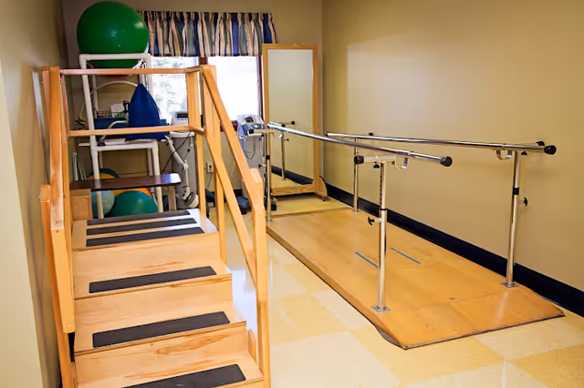 A small physical therapy room with wooden stairs equipped with handrails on both sides, a parallel bar setup for walking exercises, a full-length mirror, and shelves holding exercise balls and other therapy equipment. The room has beige walls and a window with a striped valance.