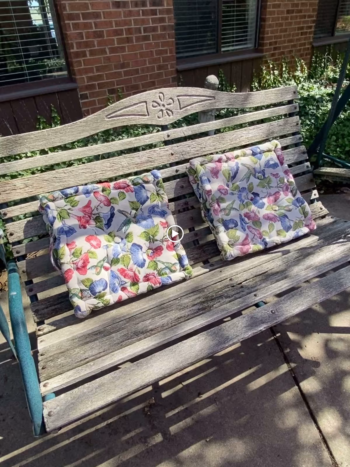 A weathered wooden outdoor bench swing with two floral-patterned cushions placed on it, situated on a concrete patio with a brick building and green plants in the background.