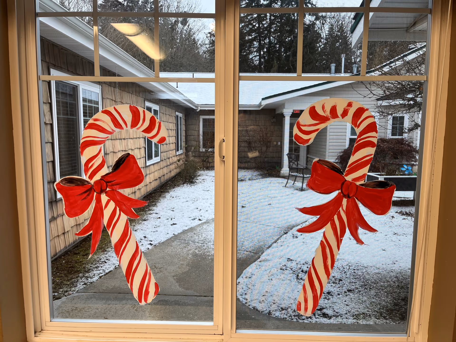 Two large painted candy canes with red bows are displayed on a window. Outside the window, there is a courtyard with light snow on the ground, surrounded by buildings with windows and some outdoor furniture.