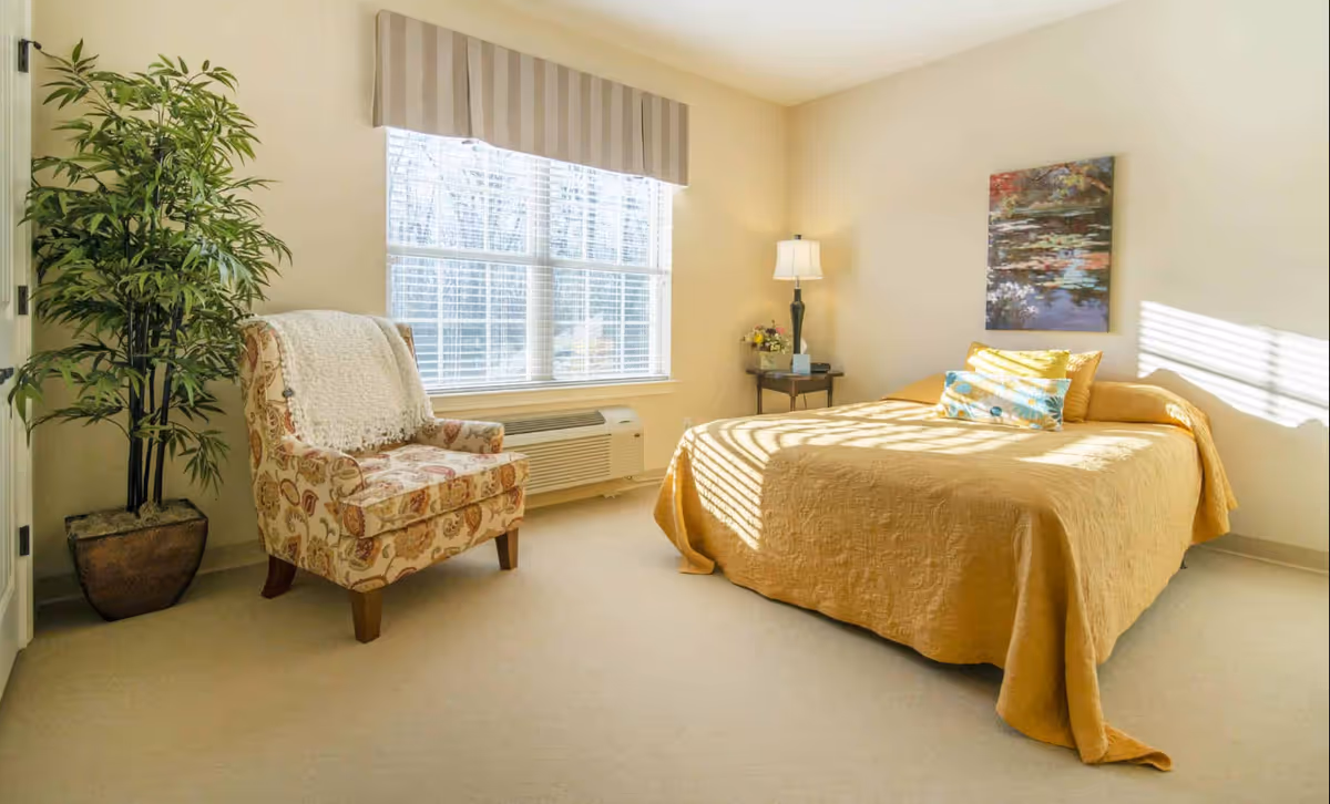 Sunny bedroom with a mustard-yellow bedspread, patterned armchair, potted plant, and a window with blinds.
