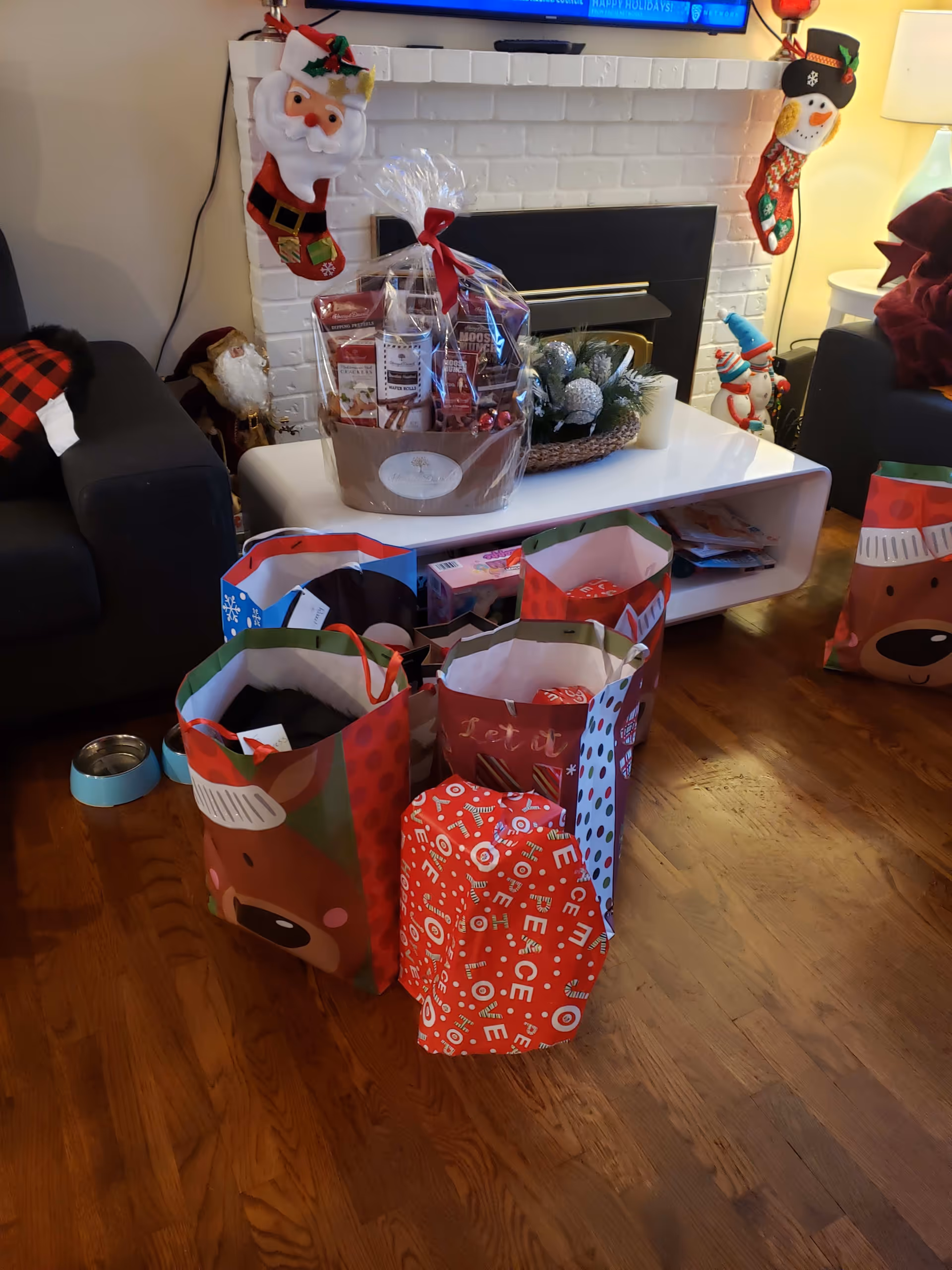 Living room with a white brick fireplace decorated for the holidays and several gift bags and presents on a wooden floor.