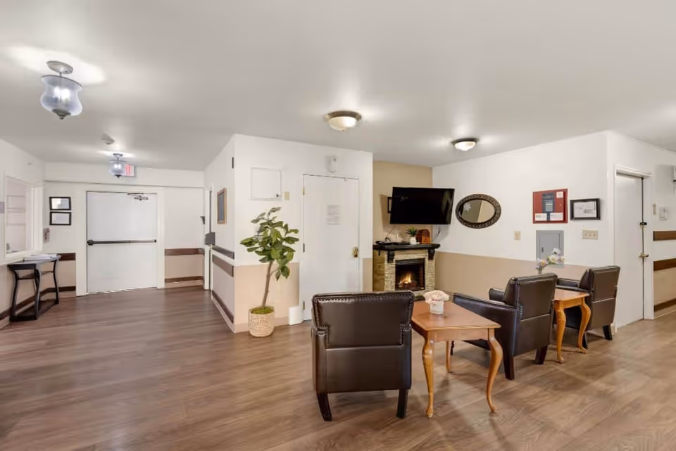 A cozy common area in a senior living facility with three dark leather armchairs arranged around two wooden side tables. A flat-screen TV is mounted above a small electric fireplace on the wall. The room has light-colored walls with a beige lower half and wood flooring. There is a potted plant near a white door, and the ceiling has multiple light fixtures. The hallway extends to the left with an exit door and some framed pictures on the walls.