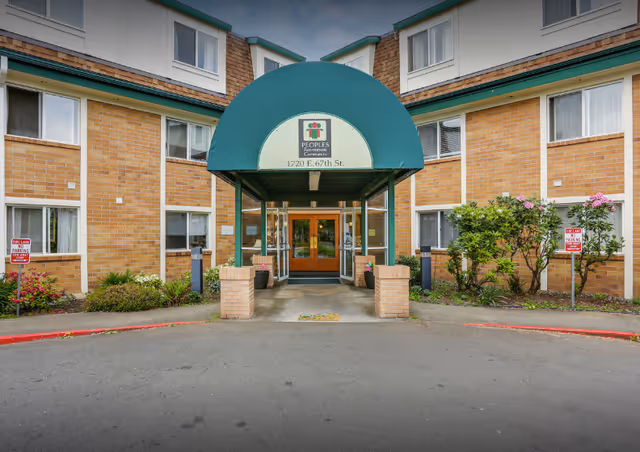 Front entrance of Peoples Senior Living facility with a green canopy above the doorway displaying the facility's name and address. The building is made of brick with multiple windows, and there are small landscaped areas with bushes and flowers on either side of the entrance.