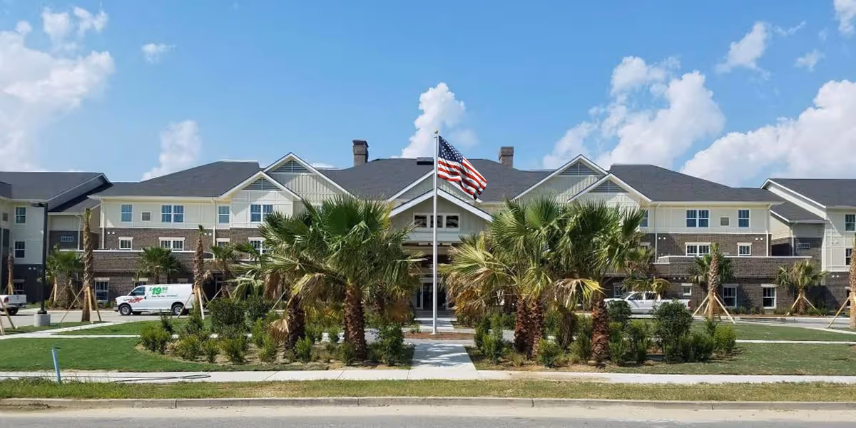 Front exterior view of Daniel Pointe Retirement Community building with a landscaped area featuring palm trees and an American flag in the center, under a partly cloudy blue sky.