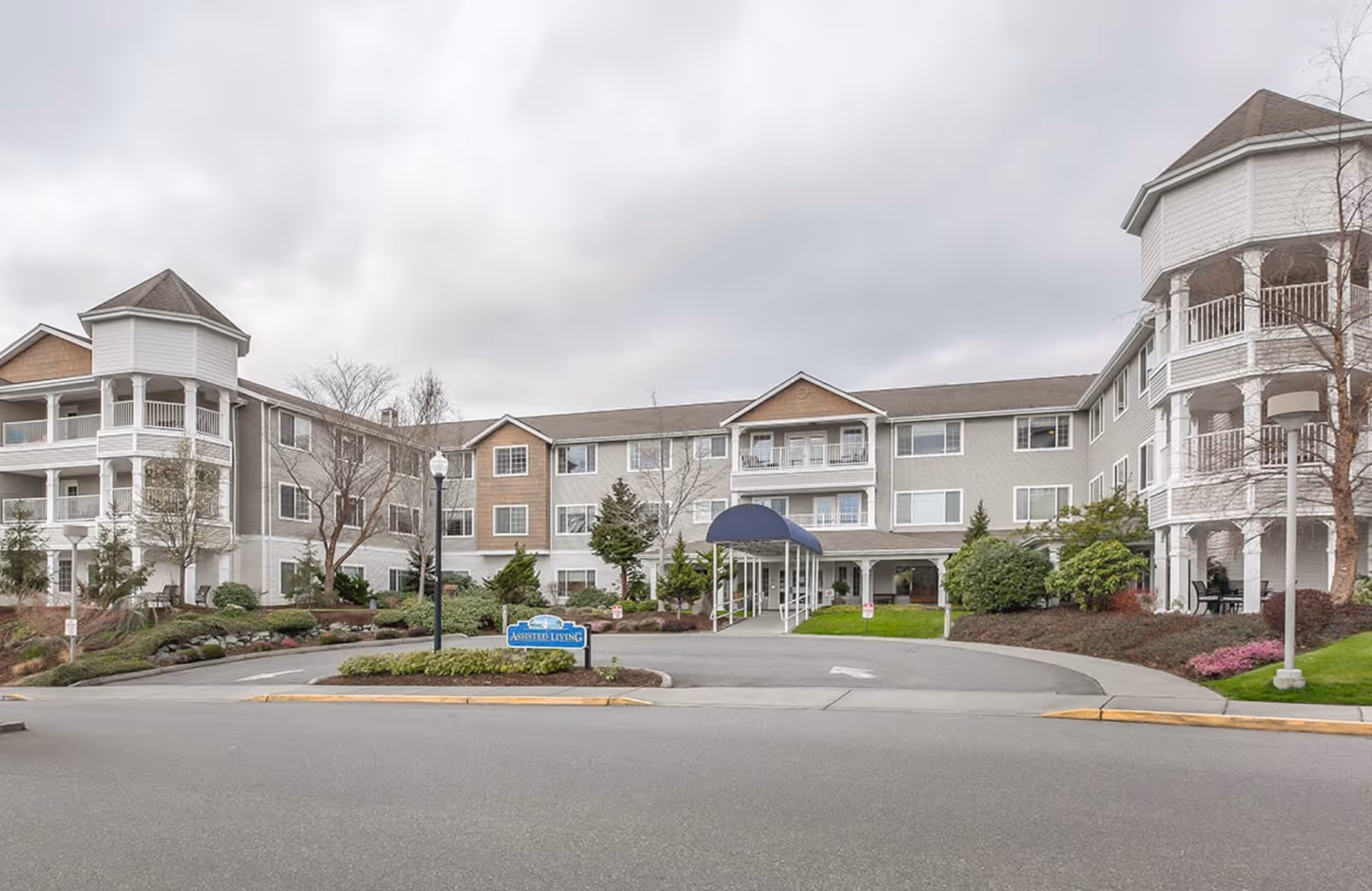 Exterior view of a three-story assisted living facility named Regency on Whidbey, featuring a central entrance with a blue canopy, landscaped greenery, and two octagonal turret-like structures on each end of the building.