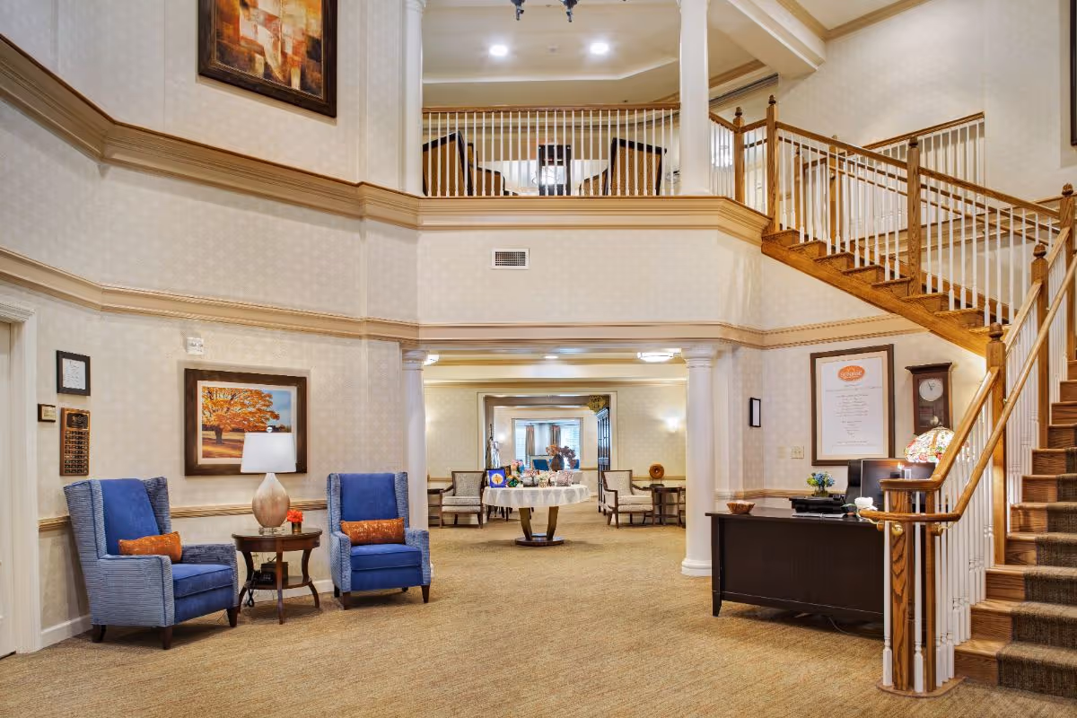 Interior view of a senior living facility lobby area with two blue armchairs and a small table with a lamp between them on the left. A staircase with wooden handrails and white balusters leads to an upper floor. There is a reception desk on the right side with a lamp and framed documents on the wall behind it. The area beyond the lobby shows more seating and a round table with decorations. The walls are light-colored with subtle patterns, and the carpet is beige.