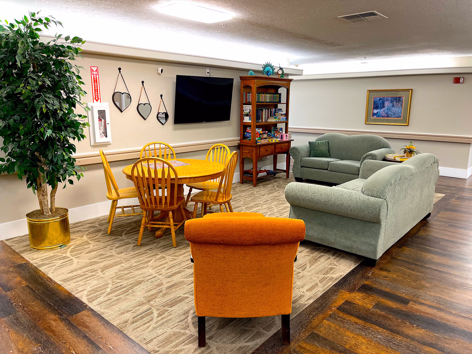 A cozy common area in an assisted living facility featuring two green upholstered sofas, an orange armchair, a wooden round table with four chairs, a wooden bookshelf with books and games, a flat-screen TV mounted on the wall, three heart-shaped mirrors, a large potted plant, and a framed picture on the wall.