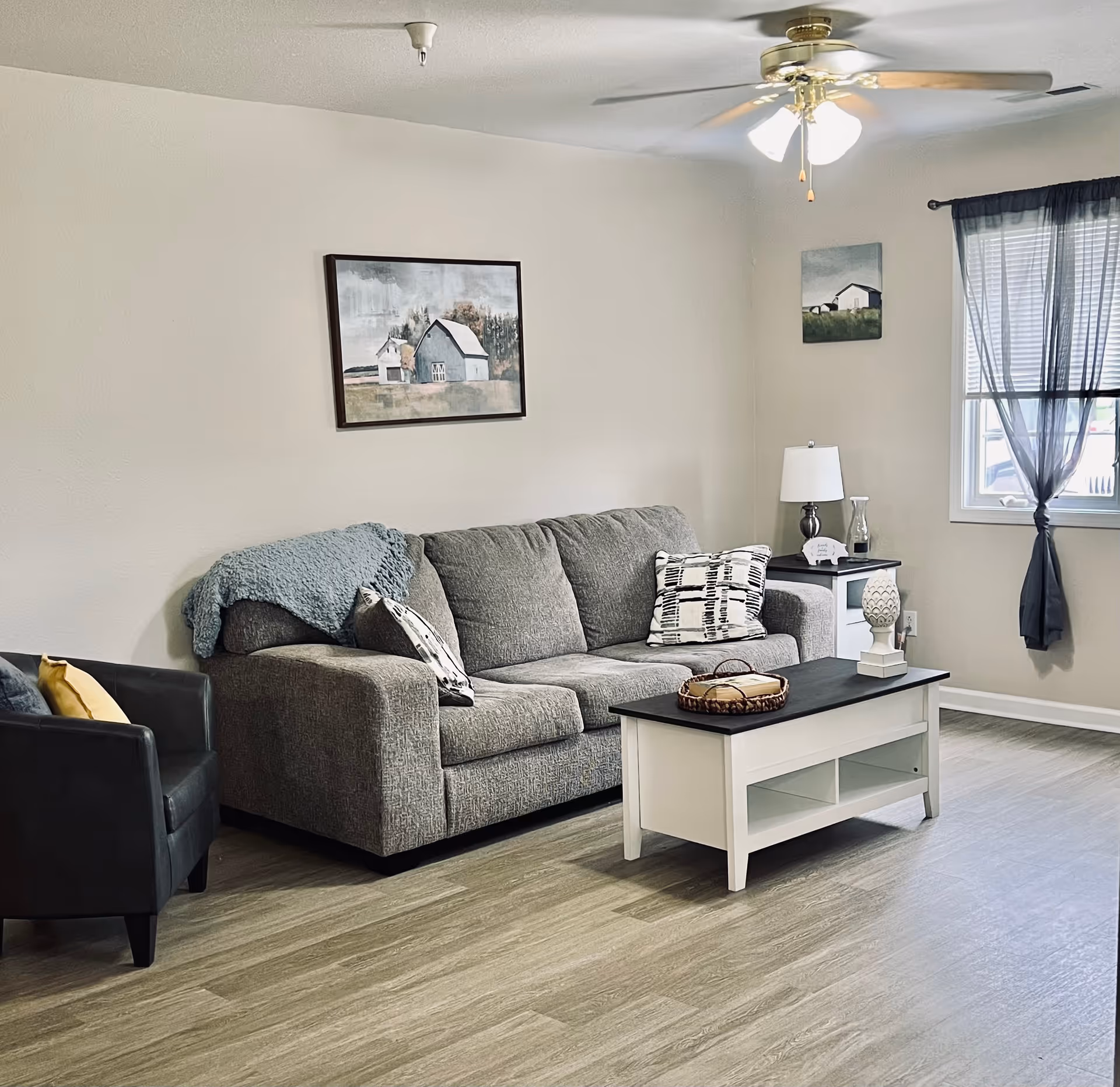 A cozy living room with a gray couch adorned with patterned pillows and a blue throw blanket. Next to the couch is a black armchair with a yellow pillow. A white coffee table with a dark top holds a decorative tray and a white ornamental piece. The room has light wood flooring, a ceiling fan with lights, and two framed paintings of barns on the walls. A window with sheer dark curtains allows natural light into the space.