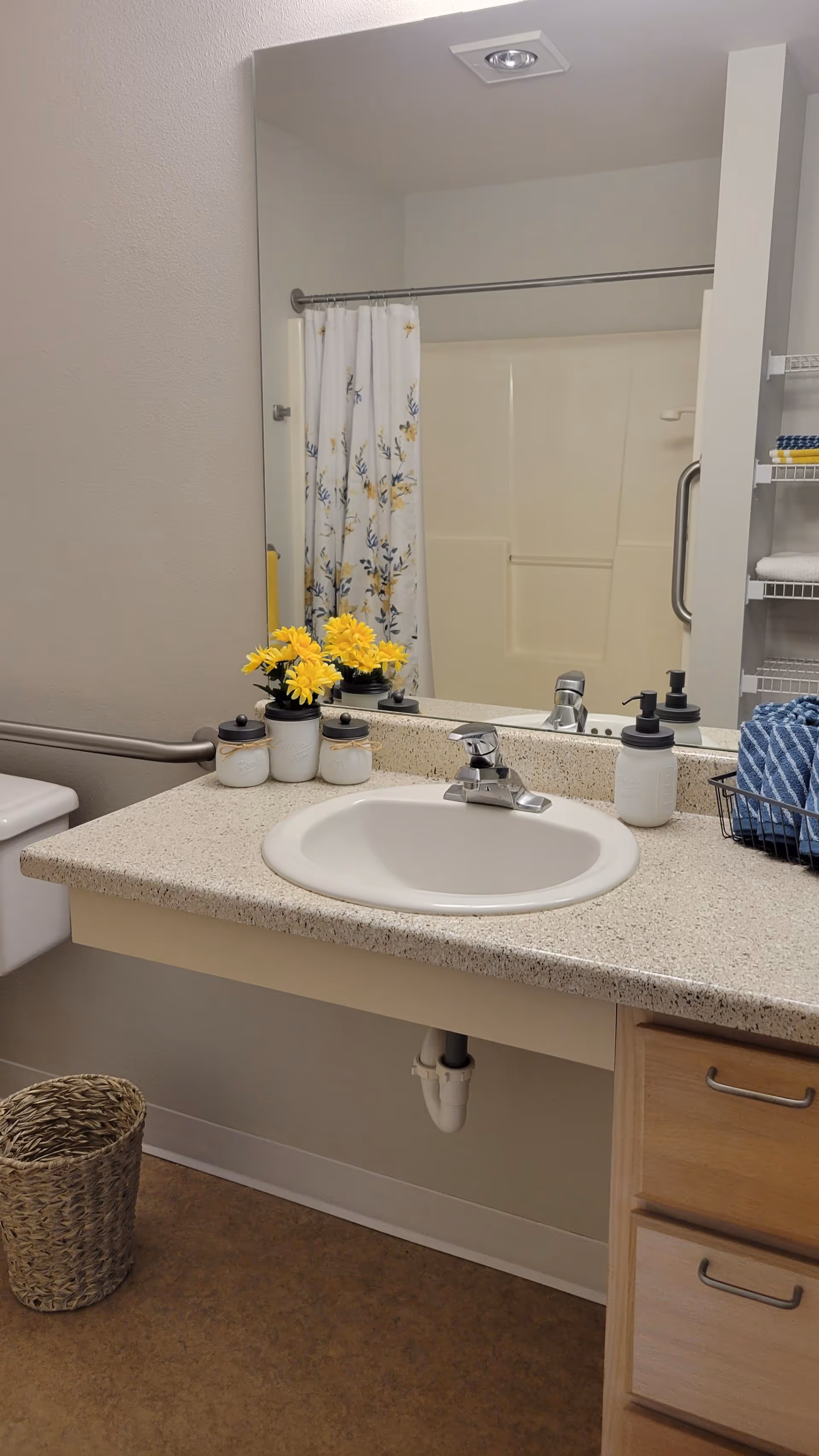 A bathroom sink with a speckled countertop, a silver faucet, and a large mirror above. On the countertop, there are three small white jars with black lids, a small vase with yellow flowers, a soap dispenser, and a wire basket holding blue towels. In the background, a shower with a white and yellow floral shower curtain is visible, along with a grab bar and shelving with folded towels. A woven wastebasket is on the floor next to the toilet.