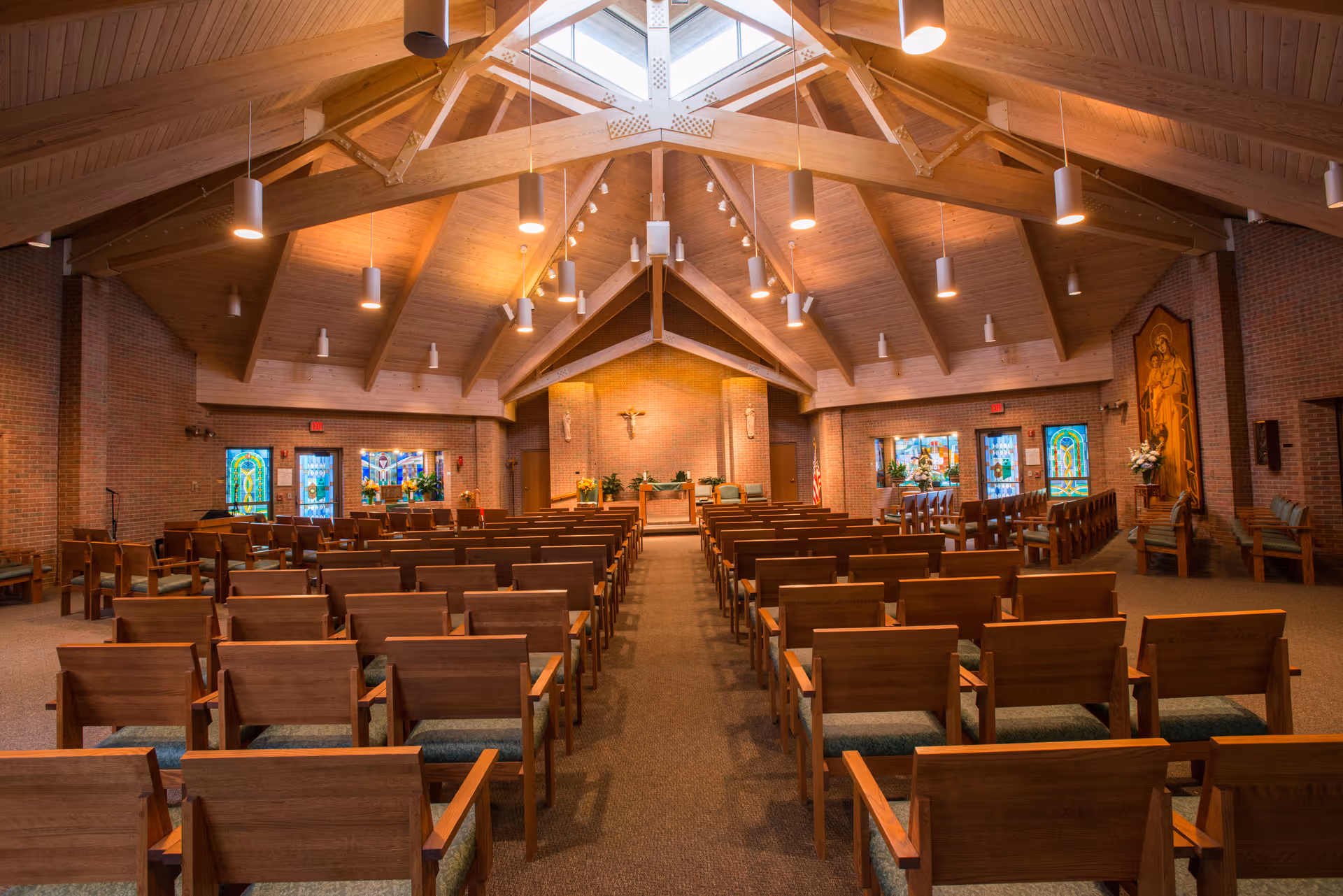 Interior view of a chapel with wooden pews arranged in rows facing an altar. The ceiling features exposed wooden beams and hanging lights. Stained glass windows and religious artwork are visible on the brick walls.