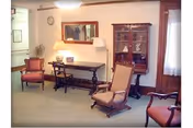 A cozy living room area with vintage wooden furniture including armchairs, a rocking chair, a long table with a lamp, and a glass-front cabinet. There is a mirror on the wall above the table and a window with curtains on the right side.