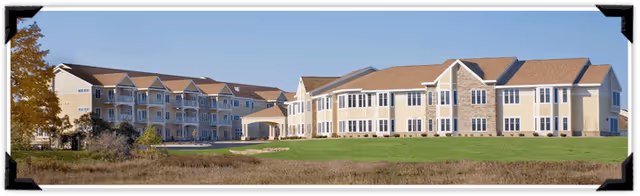 Exterior view of a large senior living facility building with beige siding and brown roofs, surrounded by green grass and a clear blue sky.