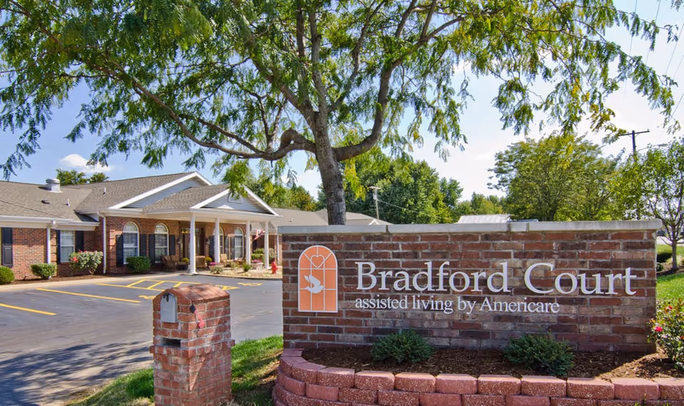 Exterior view of Bradford Court assisted living facility showing a brick sign with the facility name and a building with a covered entrance, surrounded by trees and a parking lot.