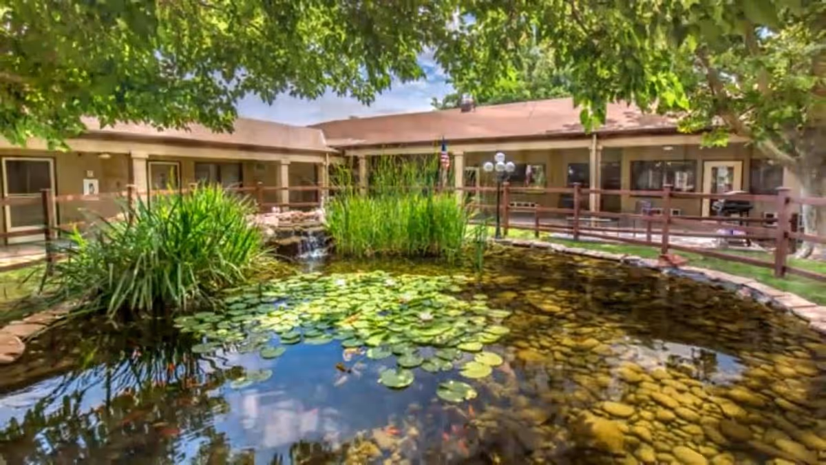 A tranquil outdoor pond with lily pads and surrounding greenery in the courtyard of a single-story assisted living facility building under a partly cloudy sky.