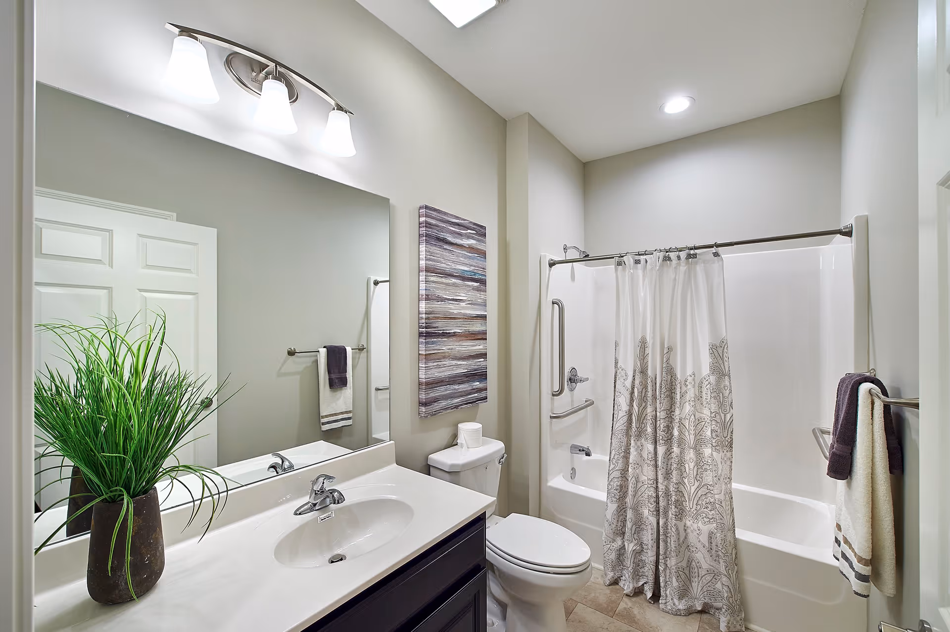 A clean and modern bathroom with a white sink and countertop, a large mirror above the sink, a potted green plant on the counter, a toilet next to the sink, and a bathtub with a patterned shower curtain. The walls are painted light gray, and there is a towel rack with towels beside the bathtub.